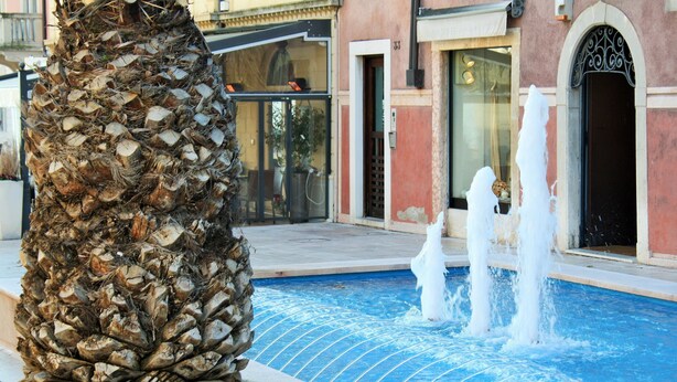 A pool with water jets, a palm tree, and a building with a number '33' in the background. - Hotel Ristorante Commercio (Salò)
