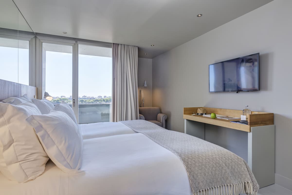 A modern hotel room with a large bed, a TV mounted on the wall, a desk with a lamp, and a view of the city through large windows.