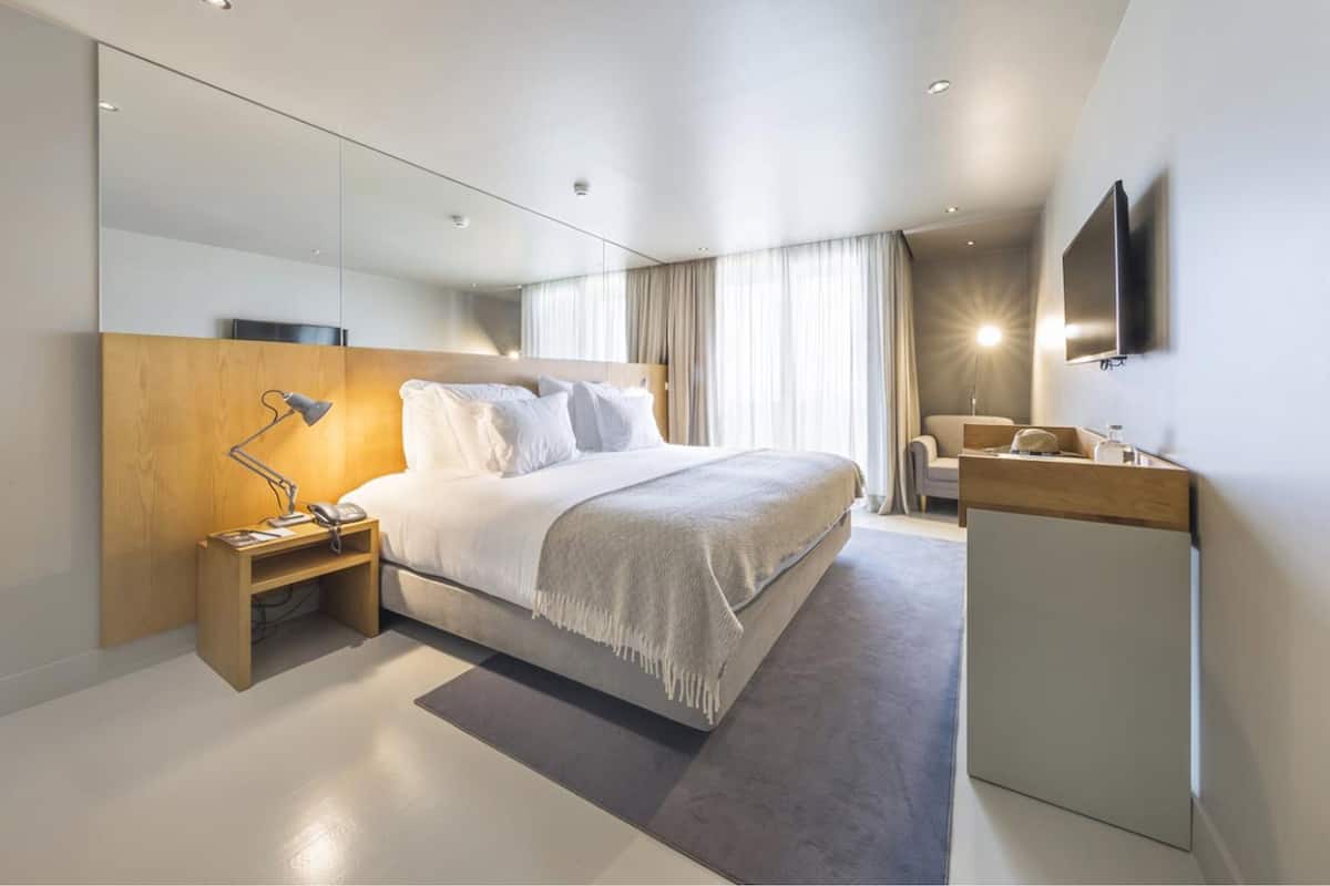 A modern hotel room with a large bed, bedside table, lamp, and a TV mounted on the wall.