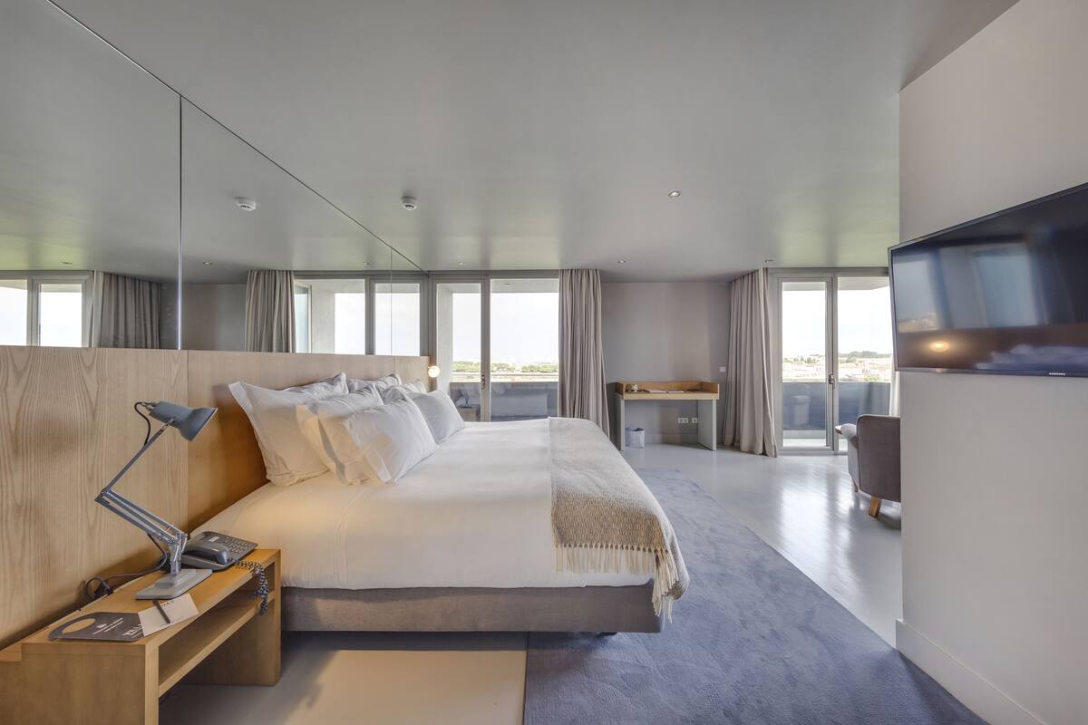 A modern hotel room with a large bed, a desk, a flat-screen TV, and a balcony with a view.