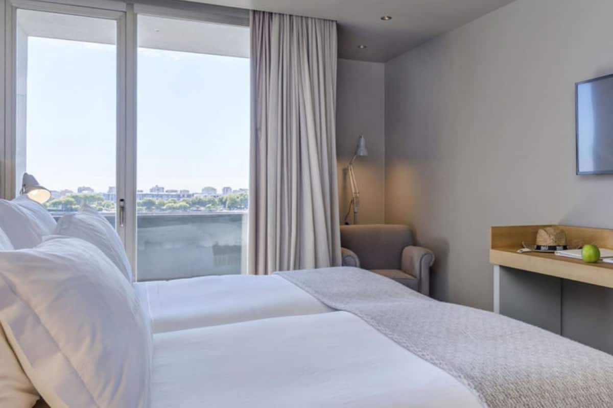 A hotel room with a large bed, a desk, a chair, a lamp, and a view of a cityscape through a window.
