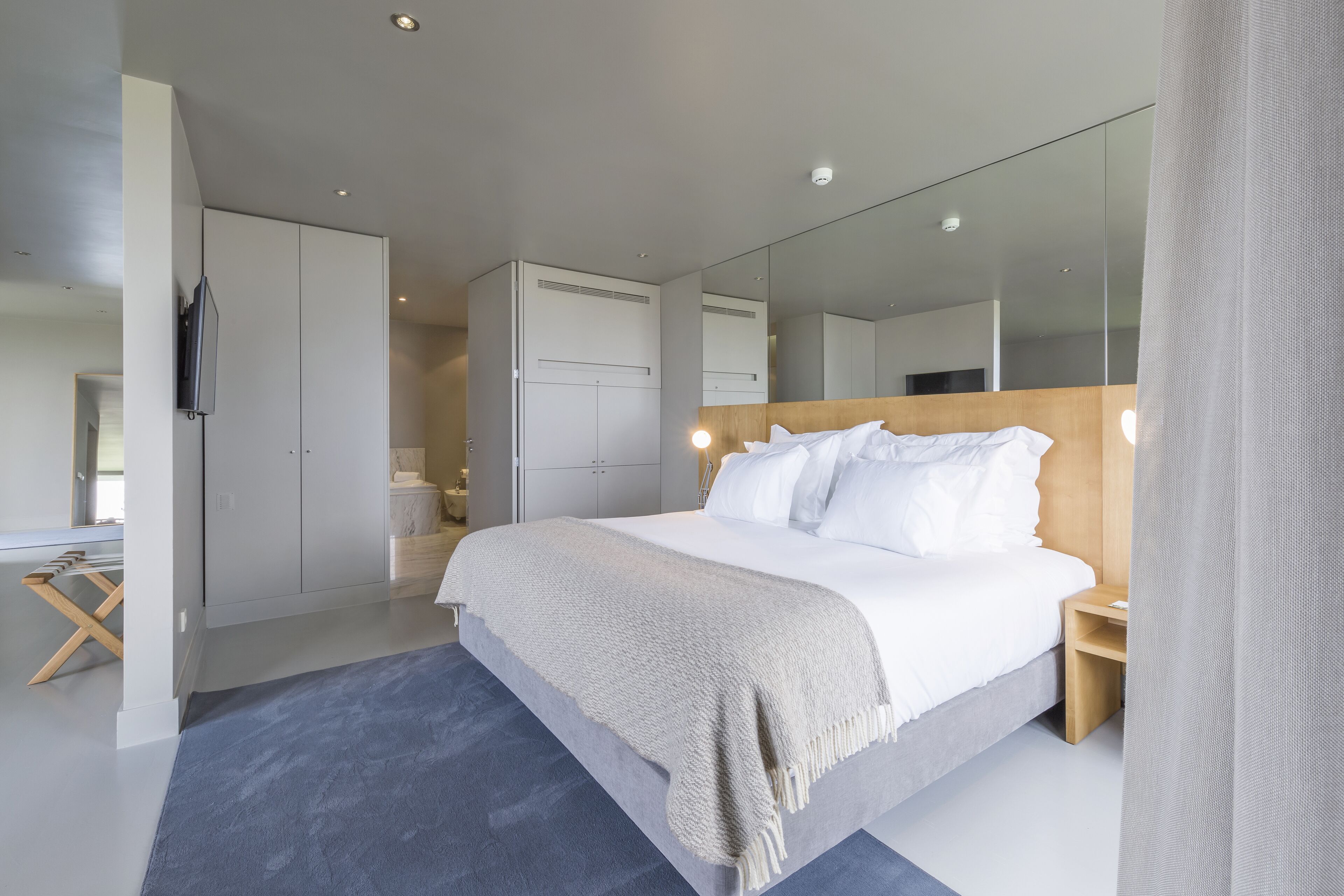 Una habitación de hotel moderna con una cama grande, cabecera de madera, mesita de noche, armario y un espejo amplio.