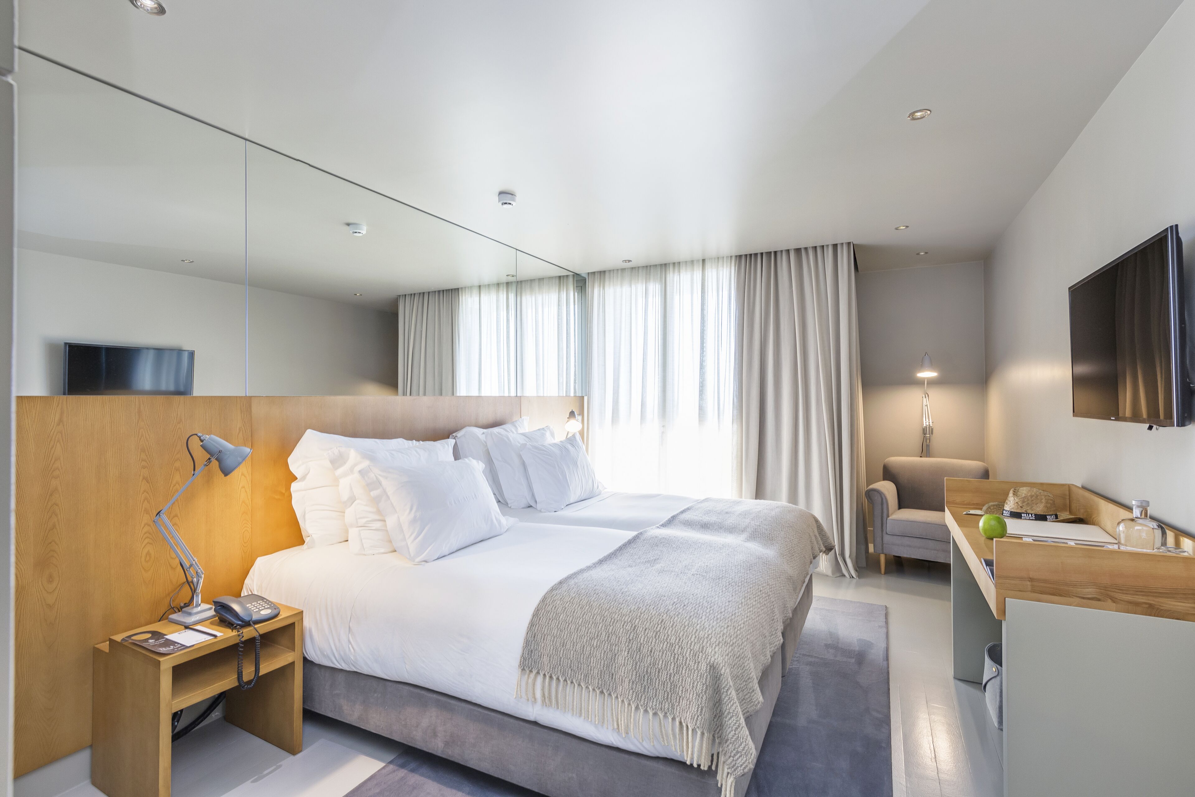 Una habitación de hotel moderna con una cama grande, una mesita de noche con lámpara, un escritorio con silla, un televisor montado en la pared y un ventanal con cortinas.