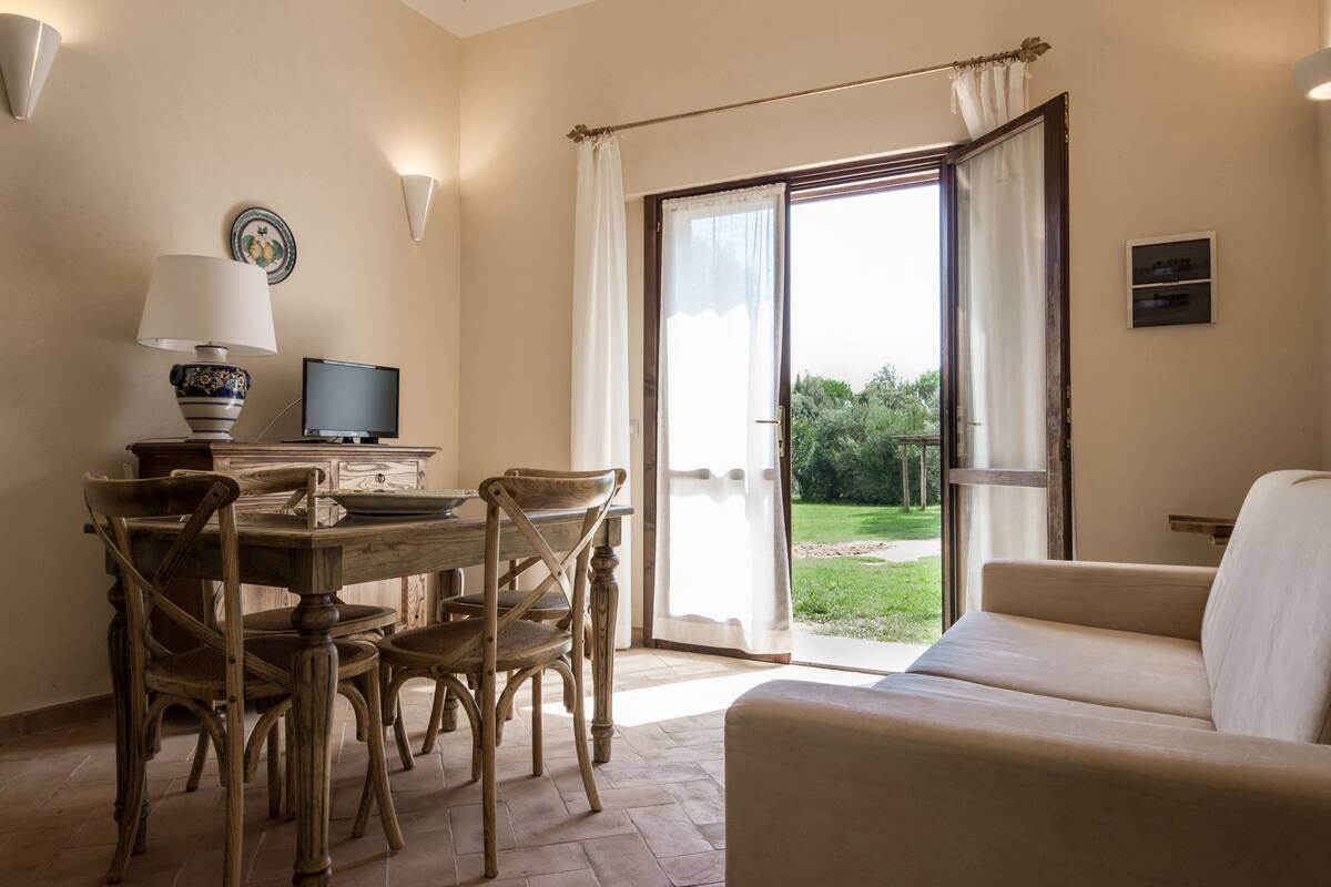 A room with a dining table, chairs, a television, a sofa, and a sliding glass door leading to a garden.