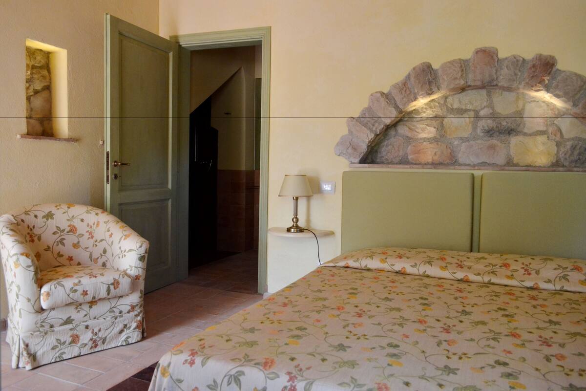 A bedroom with a floral bedspread, a chair, a stone fireplace, and a lamp.