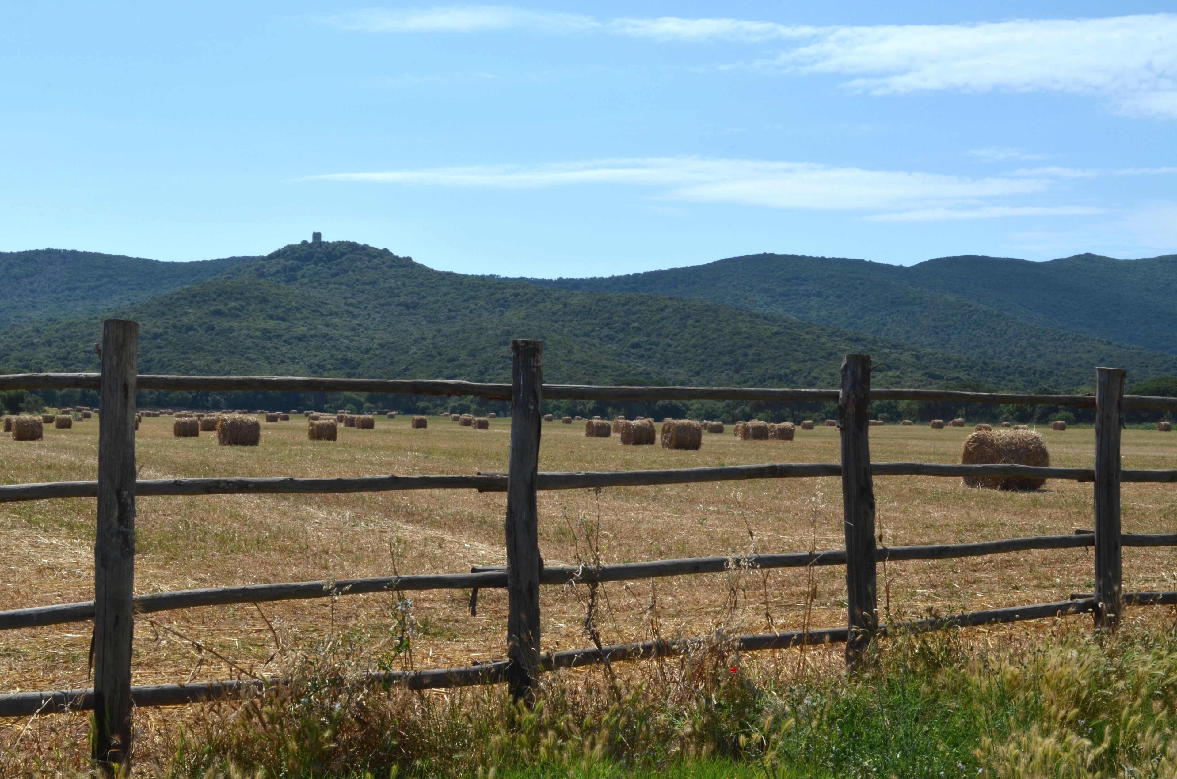 Ländliche Landschaft mit Holzzaun, Heuballen und einer Hügelkette im Hintergrund.
