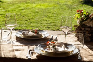 A table set for a meal with plates, cutlery, and wine glasses, set on a tablecloth with a view of a grassy area. - Tenuta Uccellina (Magliano in Toscana)
