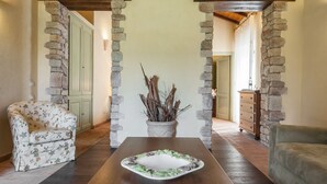 A room with a wooden table, stone pillars, a floral armchair, and a sofa. - Tenuta Uccellina (Magliano in Toscana)