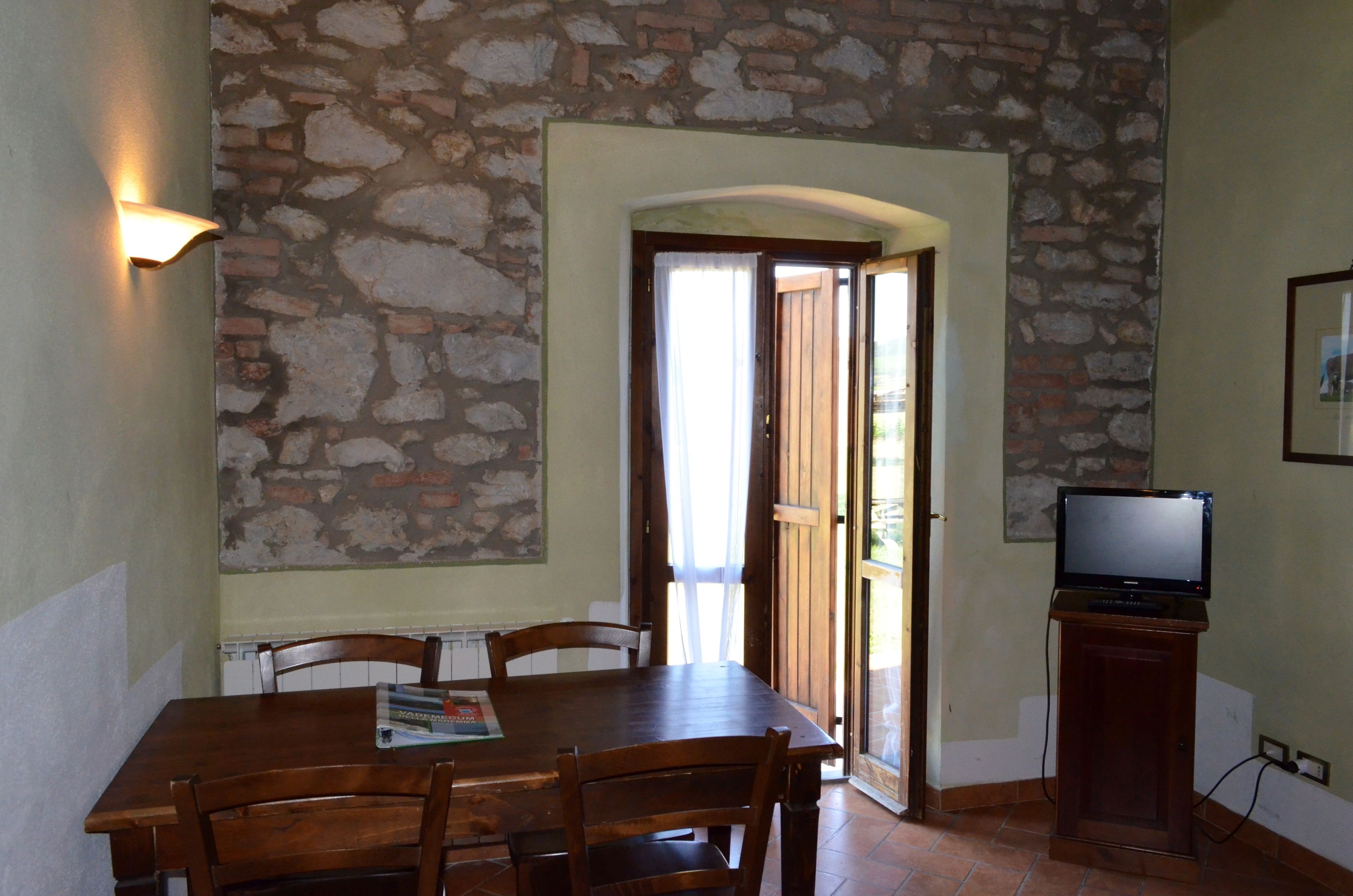 Un comedor con pared de piedra, una mesa de madera, sillas, un televisor sobre un mueble y una puerta que da al exterior.