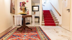 A hallway with a patterned rug, a wooden table with a vase, a mirror, and a staircase with red carpet. - Hotel Principe (Sanremo)