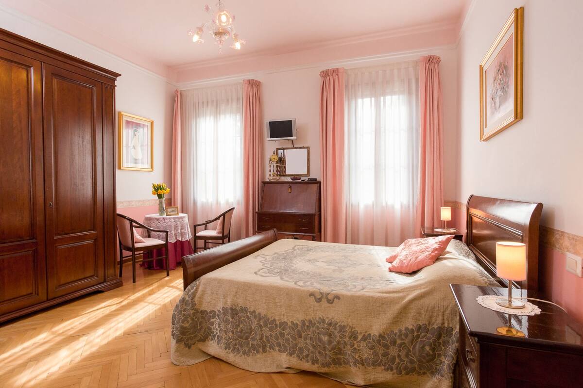 A bedroom with a bed, bedside table, lamp, dresser, desk, chair, and a small table with flowers.