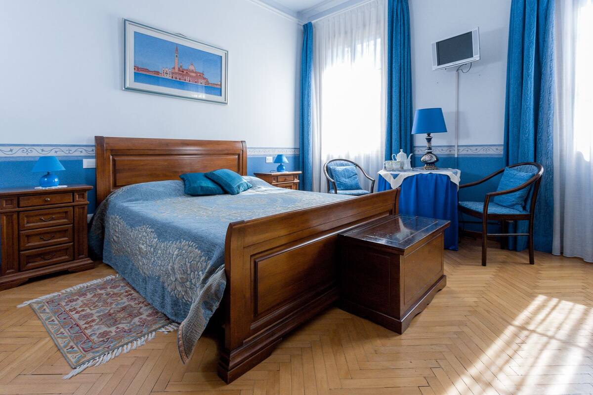 A bedroom with a wooden bed, a desk, a chair, a TV, and a window with curtains.