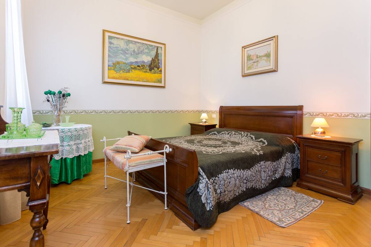 A bedroom with a wooden bed, two bedside tables with lamps, a green table with a lace tablecloth, and two framed paintings on the wall.