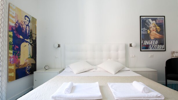 A hotel room with a bed, two pillows, a nightstand, a clock, a poster on the wall, and a chair. - Colosseum Garden (Rome)