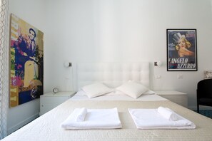A hotel room with a bed, two pillows, a nightstand, a clock, a poster on the wall, and a chair. - Colosseum Garden (Rome)
