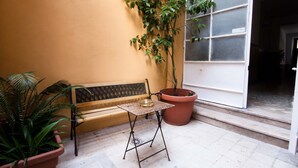 A small outdoor seating area with a bench and a table, surrounded by potted plants and a large window. - Colosseum Garden (Rome)