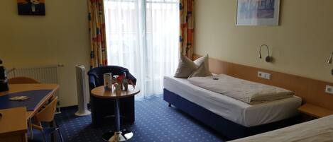 A hotel room with a bed, a desk with a chair, a small table with a bottle and a box, and a radiator.