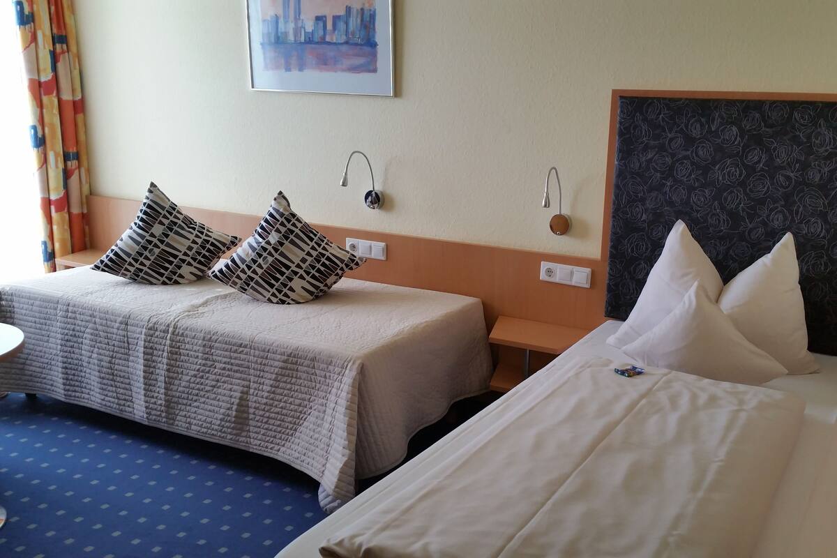 Hotellihuoneessa on kaksi sänkyä, joissa on kummassakin erilainen pääty. Huoneessa on kaksi yöpöytää, joista toisessa on lamppu ja pistorasia, sekä pieni pyöreä pöytä ja tuoli. Huoneessa on sininen, kuvioitu matto, taulu seinällä ja ikkuna, jossa on verhot.