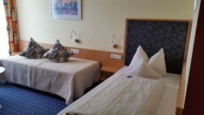 A hotel room with two beds, each with a different headboard design. There are two bedside tables, one with a lamp and a power outlet, and a small round table with a chair. The room has a blue carpet with a pattern, a painting on the wall, and a window with curtains. - Hotel Ifen (Wiesloch)