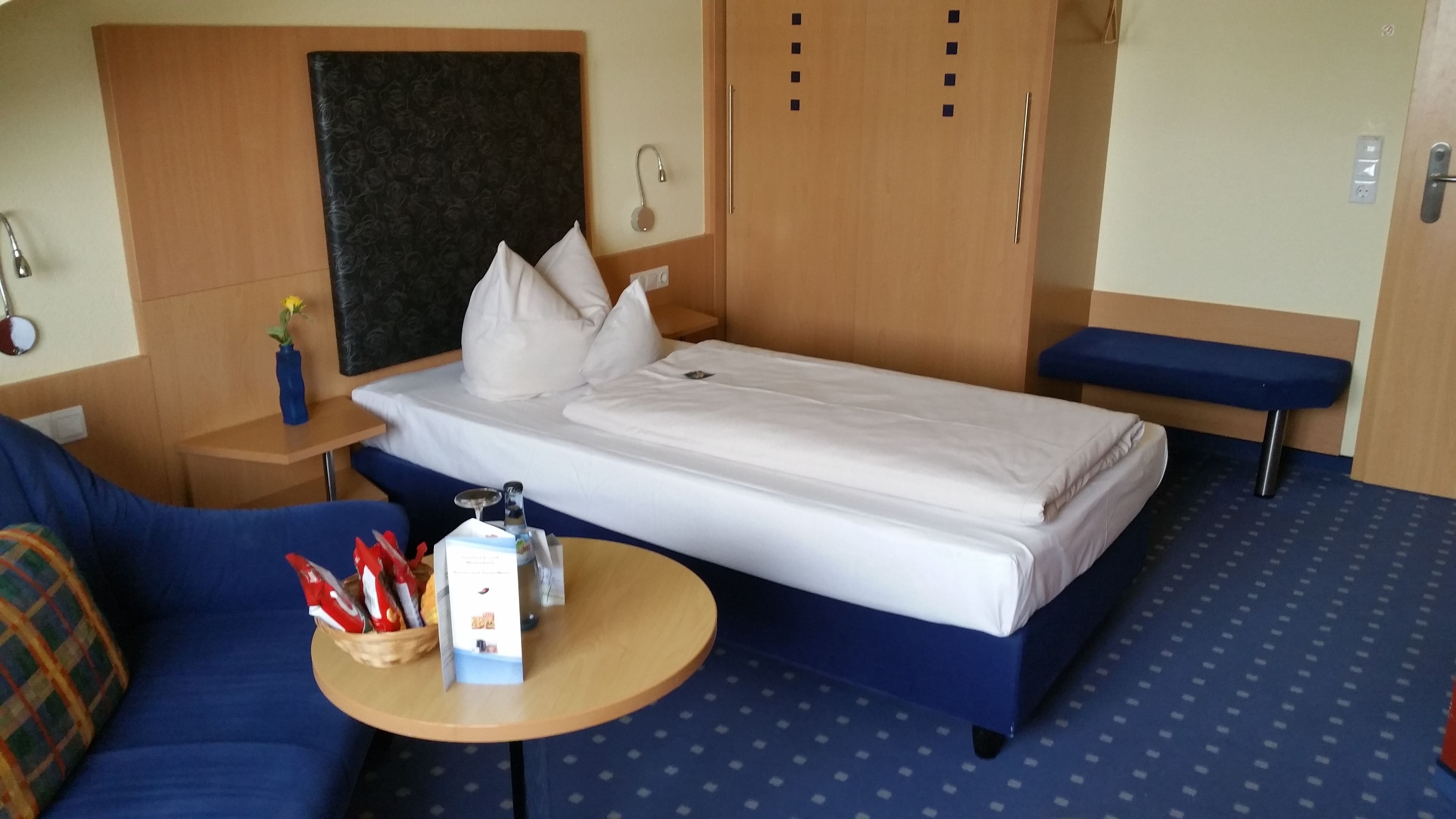 A hotel room with a bed, a small table with snacks, a bench, and a headboard.