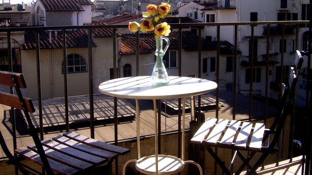 Ein Balkon mit rundem Tisch, zwei Stühlen und einer Vase mit Blumen. Im Hintergrund ist ein kuppelförmiges Gebäude zu sehen.