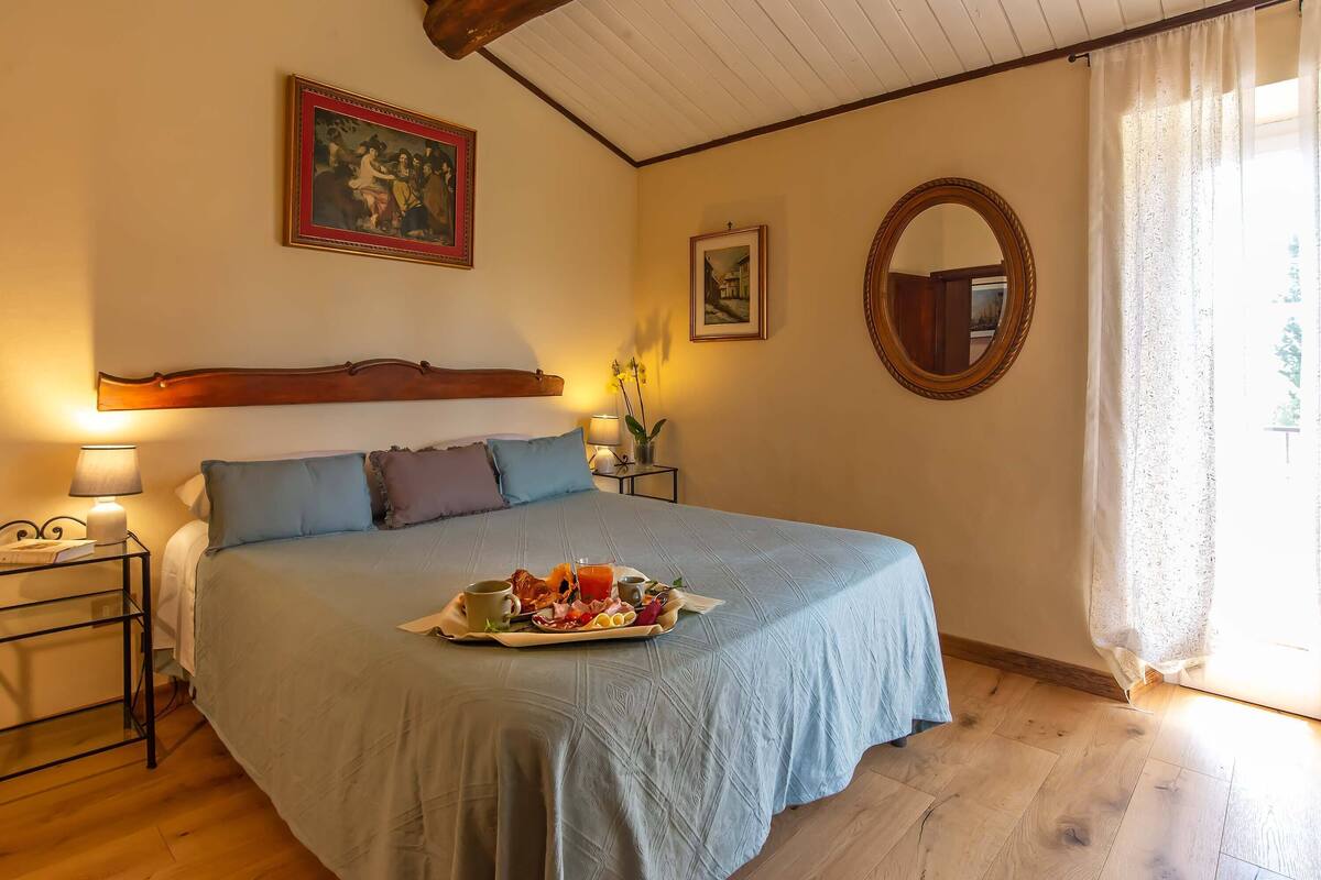 A bedroom with a bed, a bedside table with a lamp, a tray with breakfast items, and a window with curtains.