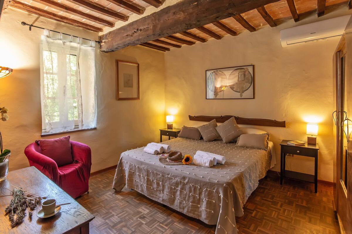 A cozy bedroom with a large bed, two bedside tables with lamps, a red armchair, and a wooden floor.