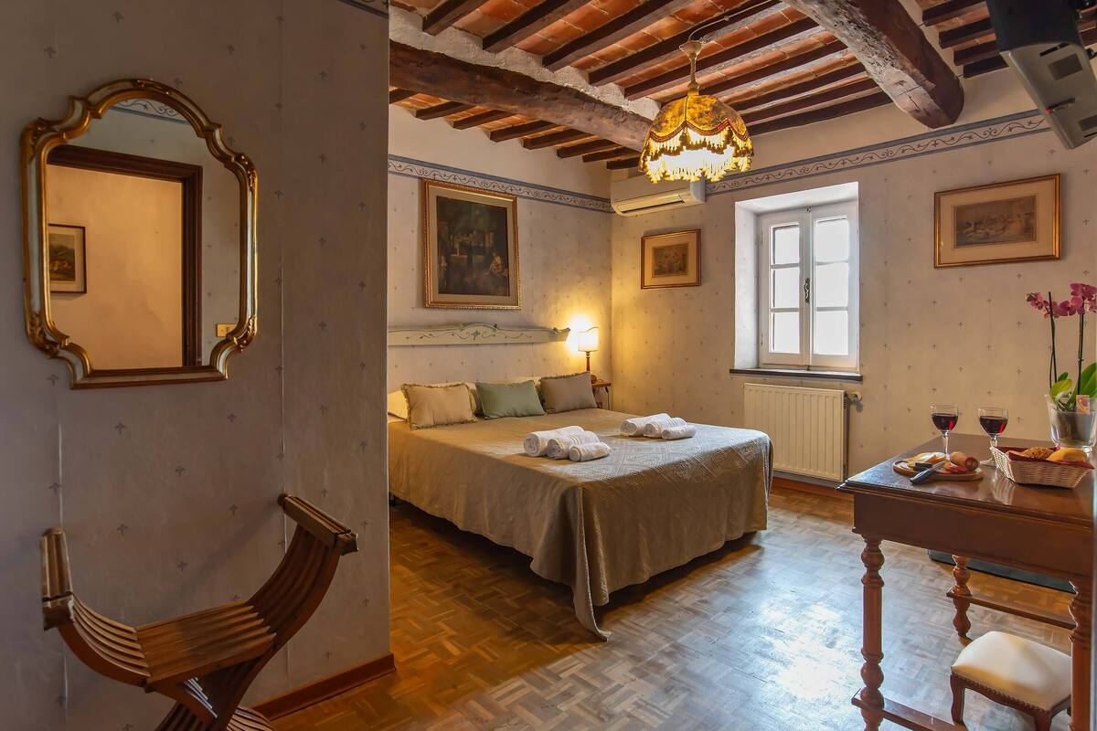 A bedroom with a bed, a wooden chair, a table with a vase, and a window.