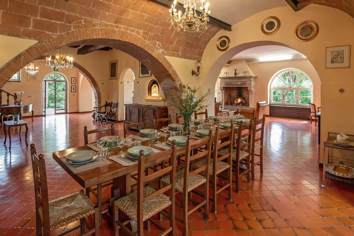 A spacious dining area with a long wooden table set for a meal, surrounded by brick arches and a chandelier.