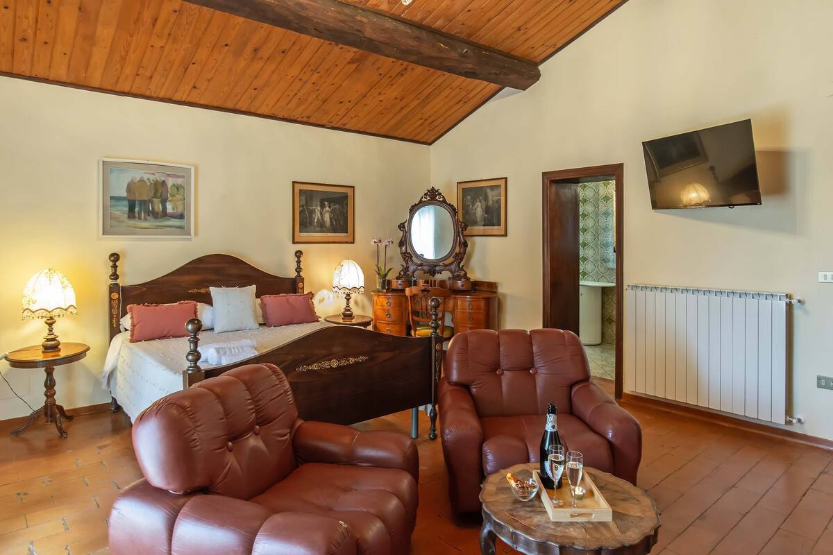 A hotel room with a wooden ceiling, a bed with a headboard, two brown leather chairs, a wooden side table with a bottle and glasses, and a TV mounted on the wall.