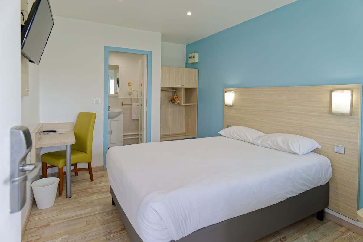 Quarto de hotel com cama, escrivaninha, cadeira, televisão e banheiro visível através de uma porta aberta.
