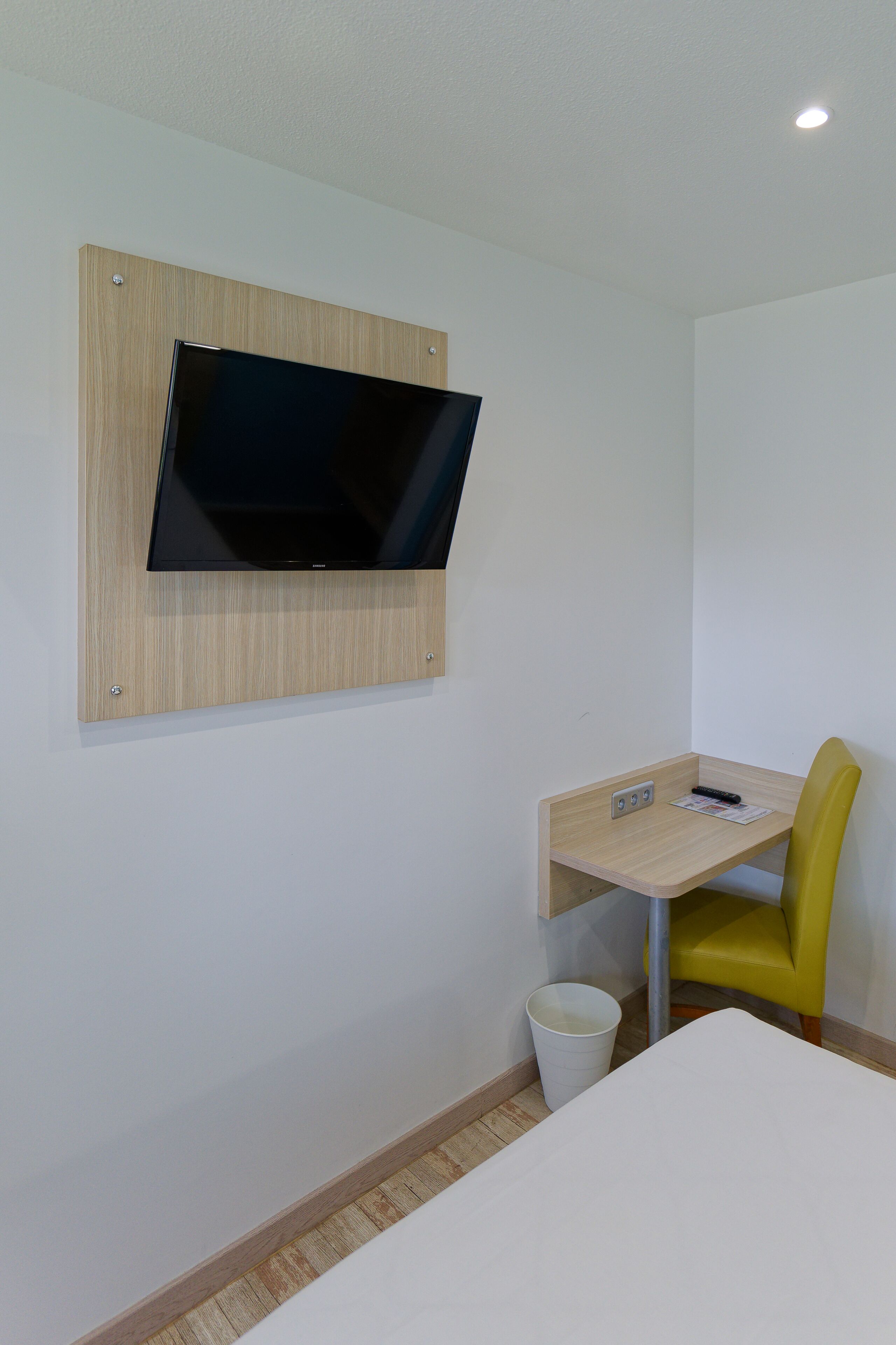 A hotel room with a mounted TV, a small desk with a chair, and a trash bin.