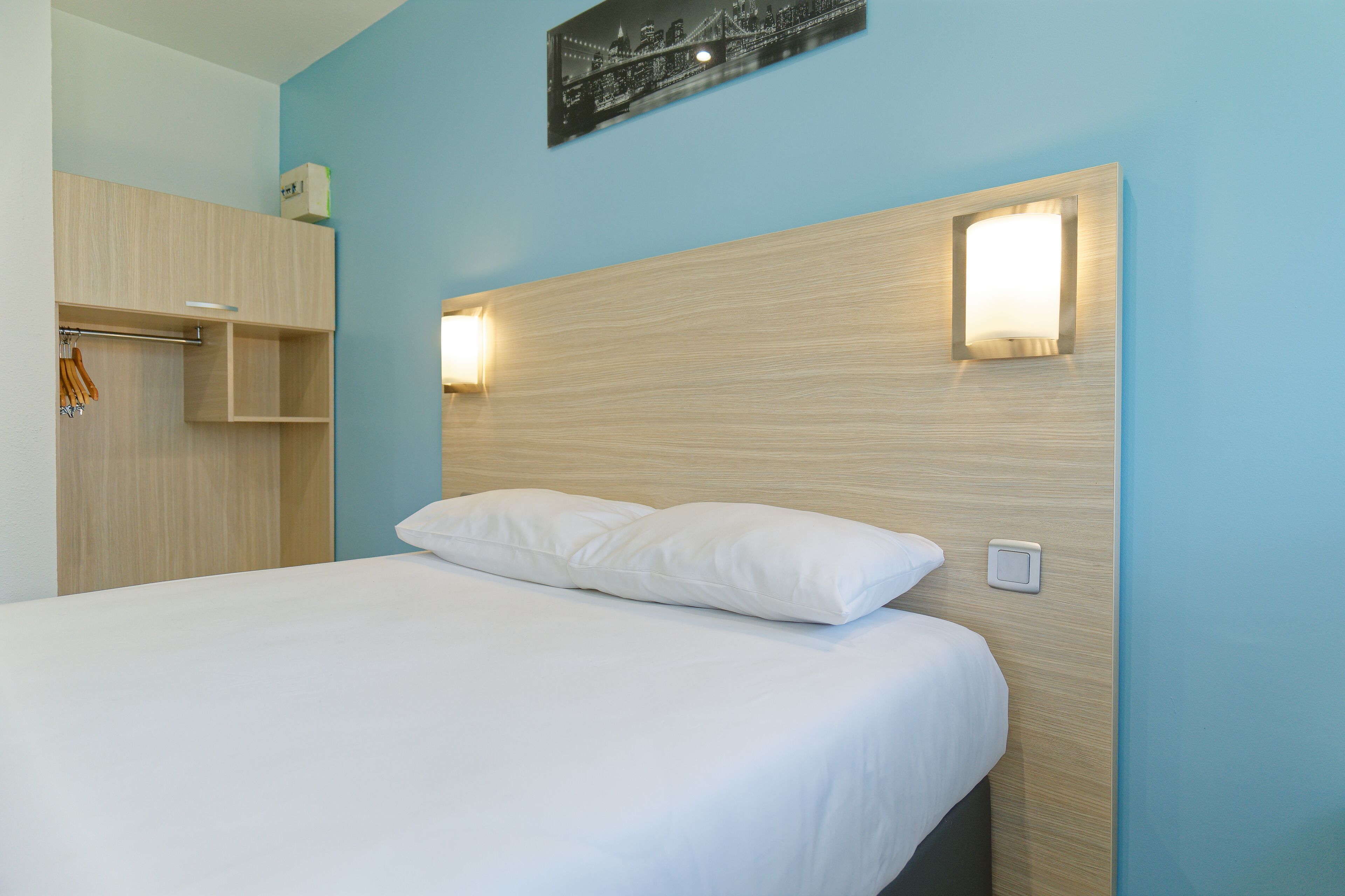 Une chambre d’hôtel avec un lit, une tête de lit en bois, deux lampes fixées au mur et une œuvre d’art représentant un paysage urbain au-dessus du lit.