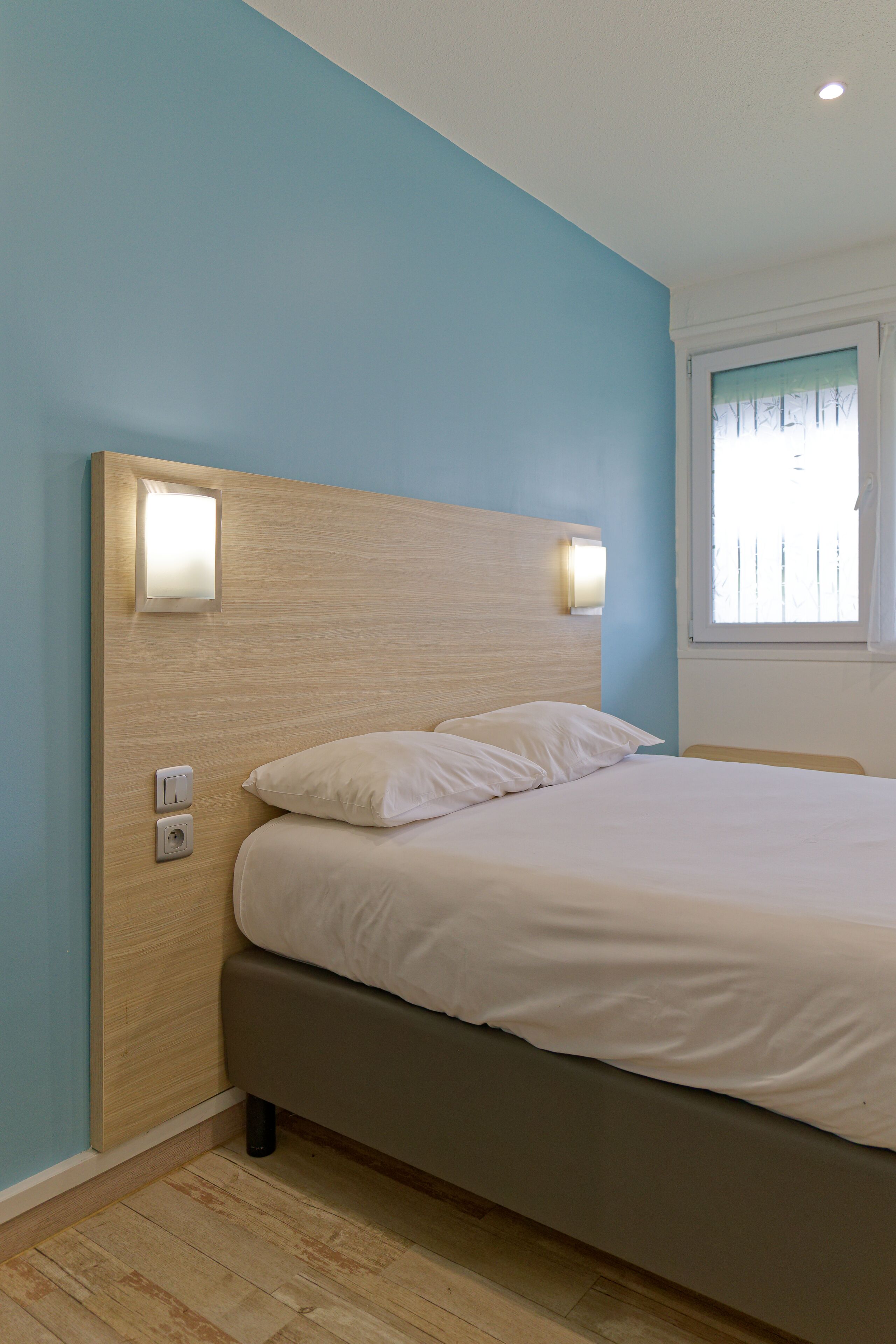 A bedroom with a bed, a headboard with wall-mounted lights, a window with blinds, and a wooden floor.