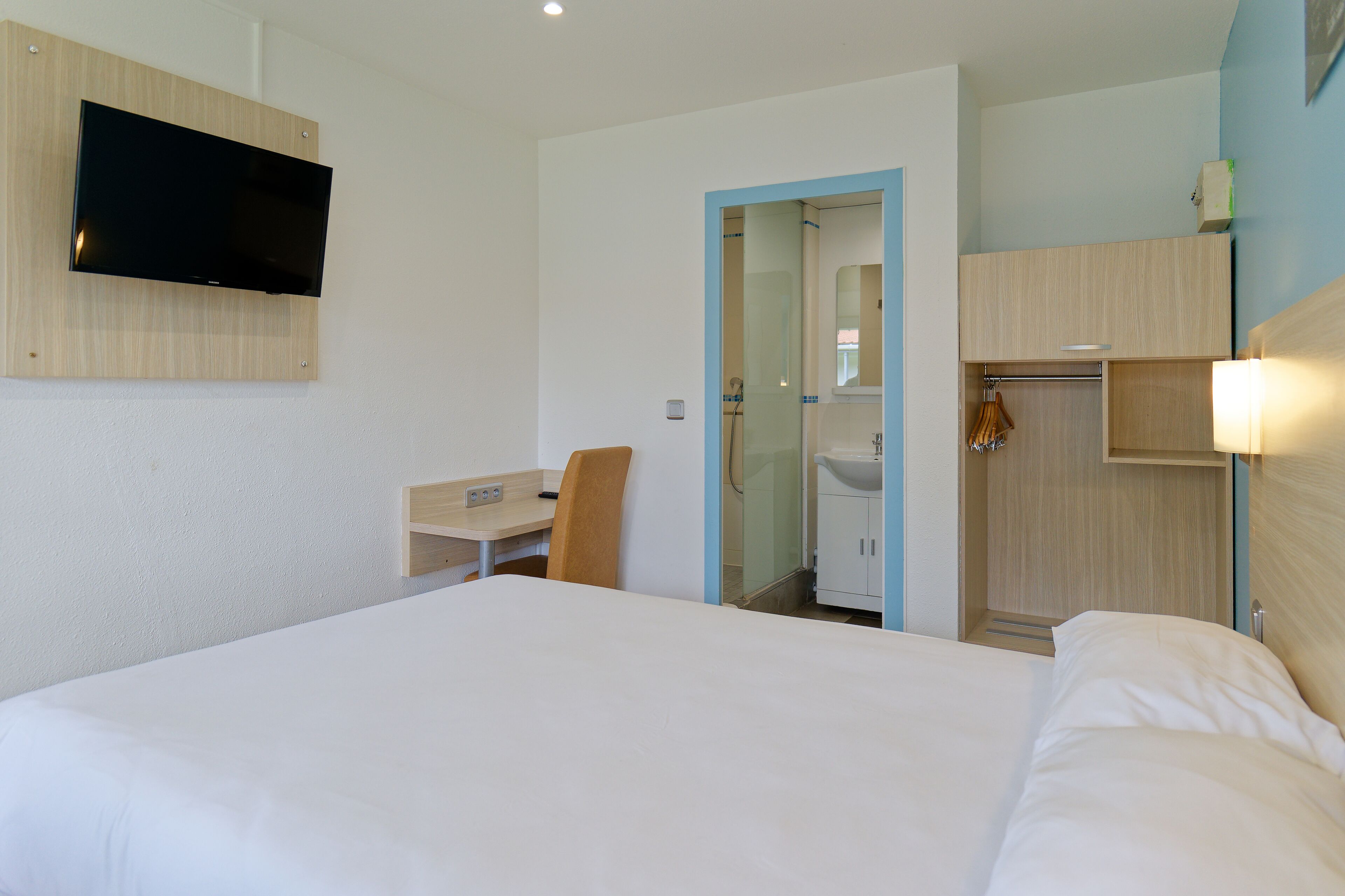A hotel room with a bed, a desk, a chair, a TV mounted on the wall, and a bathroom visible through an open door.