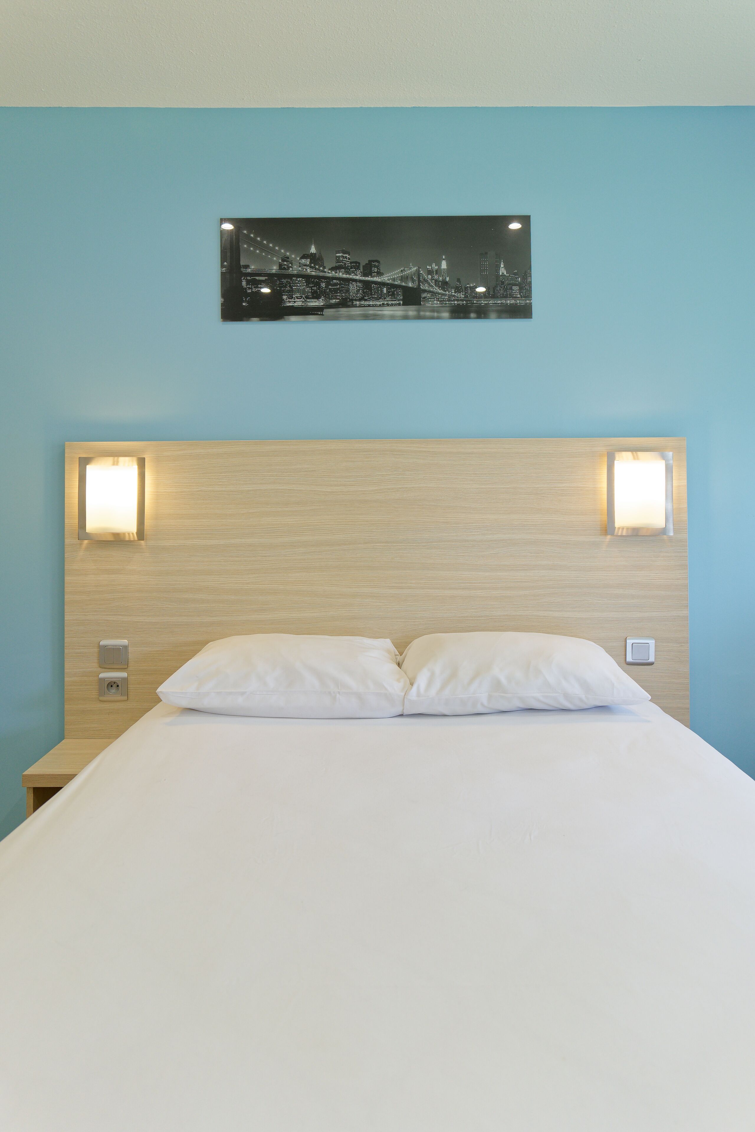 A neatly made bed with white linens, a wooden headboard, and two wall-mounted lights.