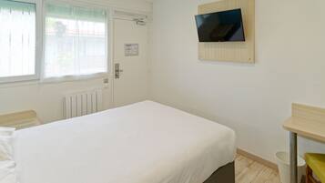 A bedroom with a bed, a television mounted on the wall, a window with blinds, and a radiator.