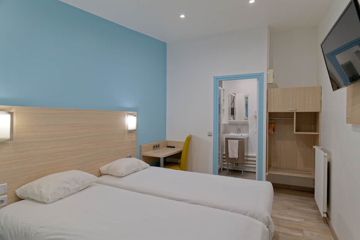 Quarto de hotel com duas camas, uma escrivaninha, uma cadeira, banheiro e televisão presa na parede.