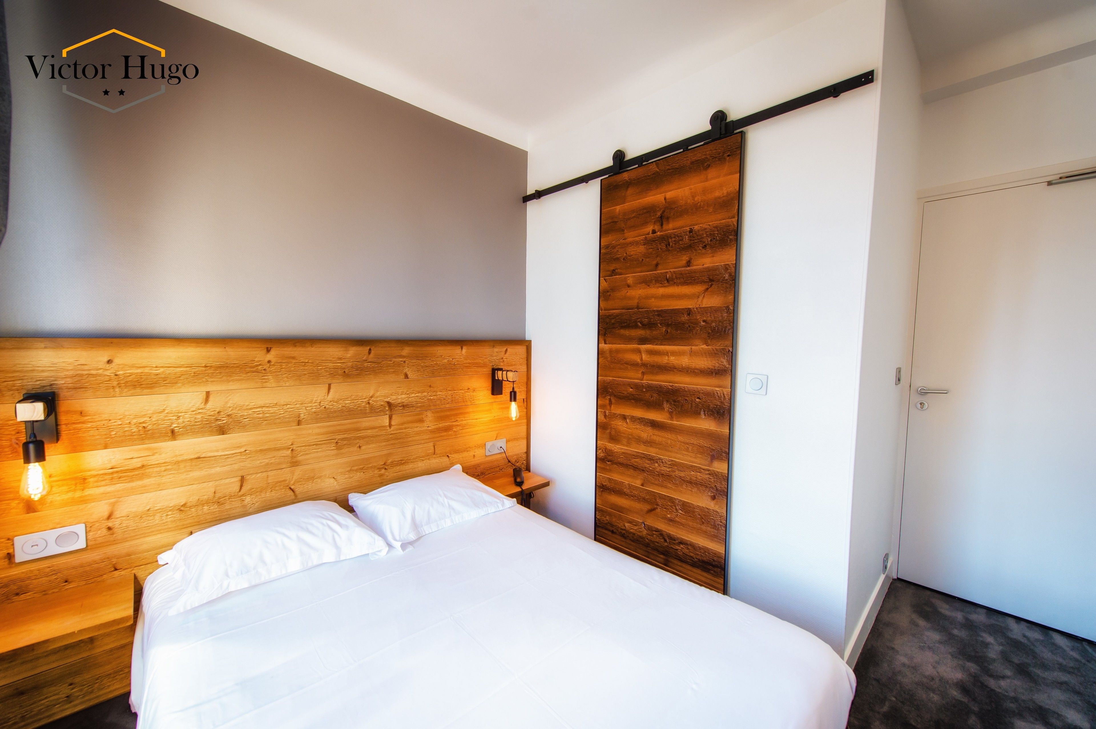 A hotel room with a wooden headboard, a bed with white linens, a nightstand with a lamp, and a sliding door.