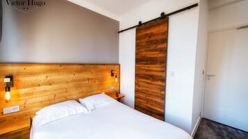 Habitación de hotel con cabecera de madera, cama con ropa blanca, mesita de noche con lámpara y puerta corredera.