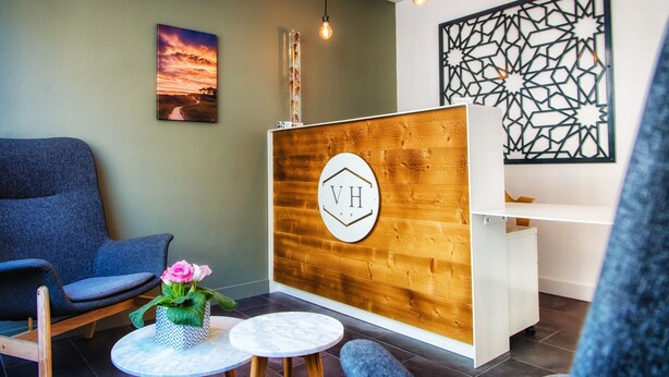 A reception area with a wooden reception desk, a blue chair, a small table with a vase of flowers, and a wall-mounted sign with 'VH'. - Victor Hugo (Lorient)