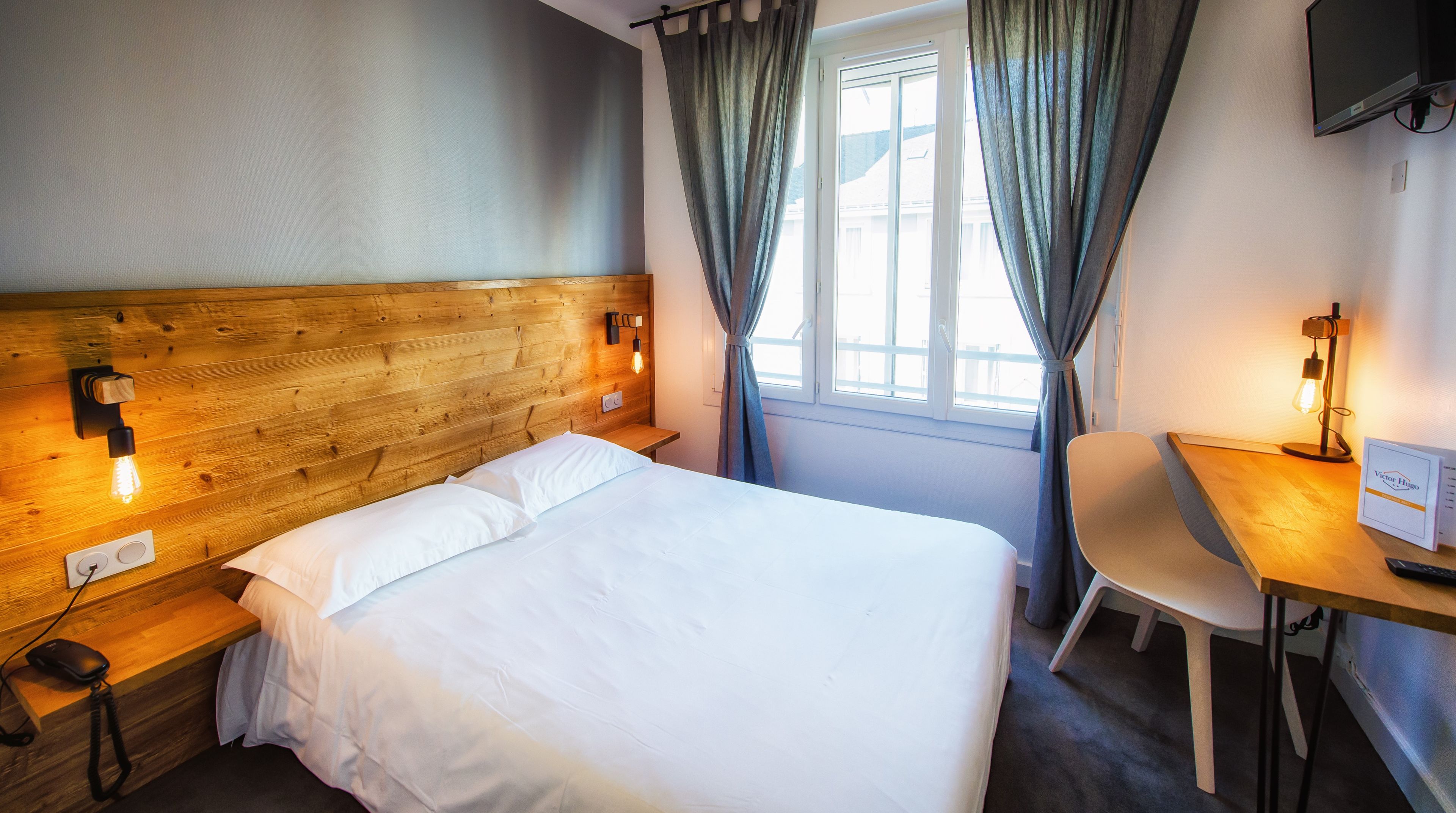 Ein Hotelzimmer mit hölzernem Kopfteil, einem Bett mit weißer Bettwäsche, einem Nachttisch mit Lampe, einem Sessel und einem Schreibtisch mit Schild.
