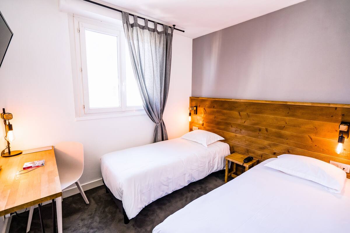 Habitación de hotel con dos camas individuales, un cabecero de madera, un escritorio con silla, una ventana con cortinas y una mesita de noche con lámpara.