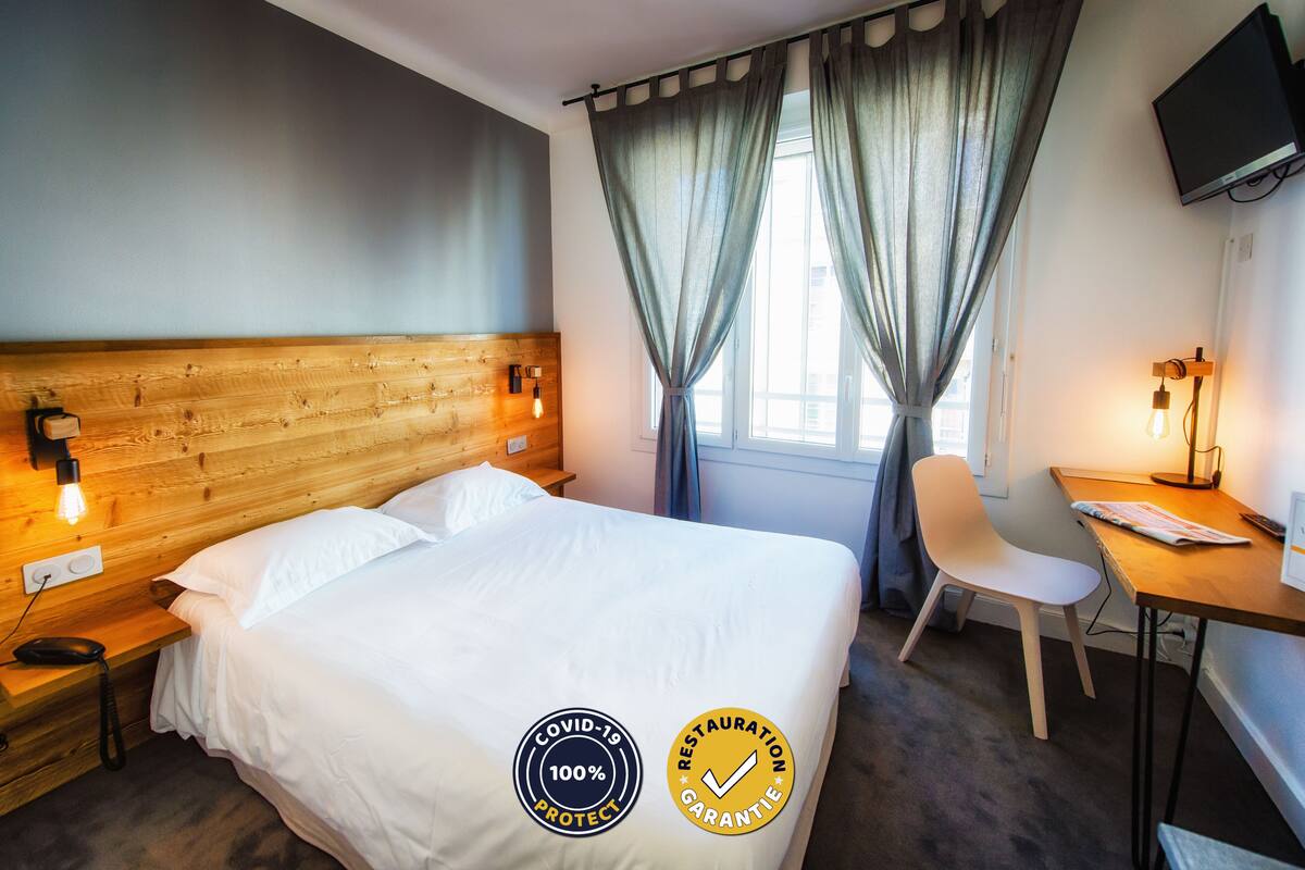 Habitación de hotel con cabecera de madera, cama con ropa blanca, escritorio con silla y televisor montado en la pared.