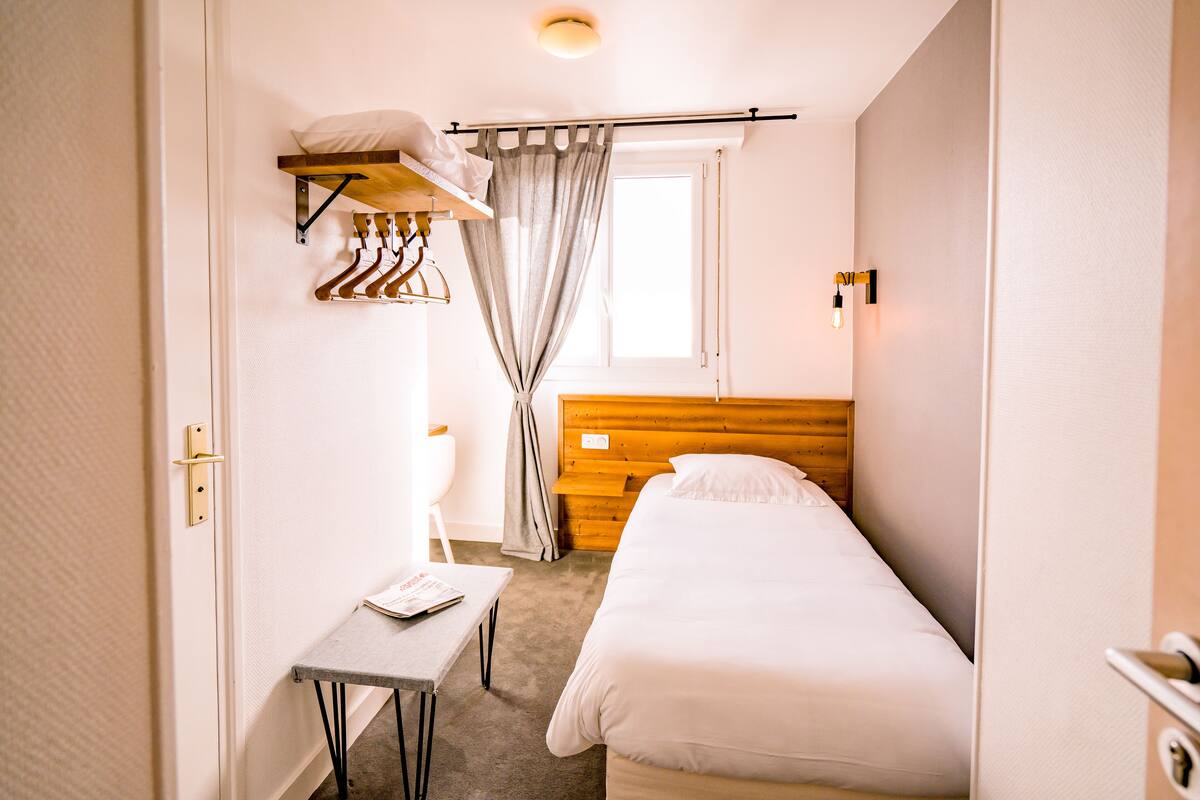 Habitación de hotel compacta con una cama individual, una mesita pequeña, un perchero y una ventana con cortinas.