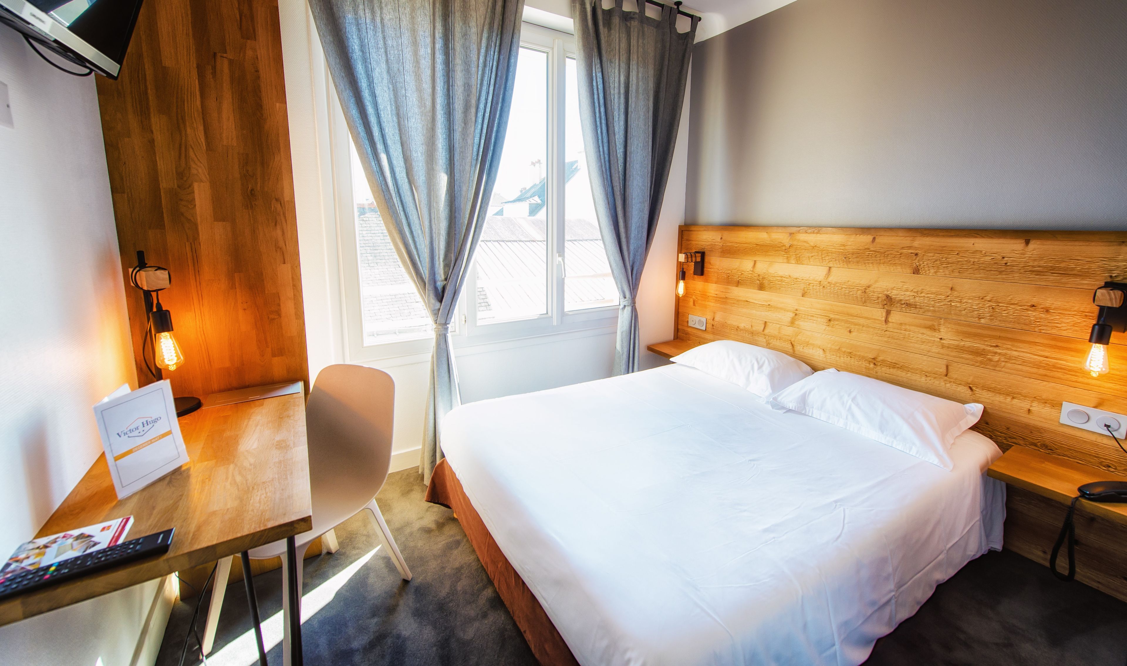 A hotel room with a wooden headboard, a bed with white linens, a desk with a chair, a lamp, and a window with curtains.