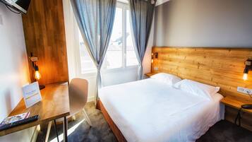 A hotel room with a wooden headboard, a bed with white linens, a desk with a chair, a lamp, and a window with curtains.