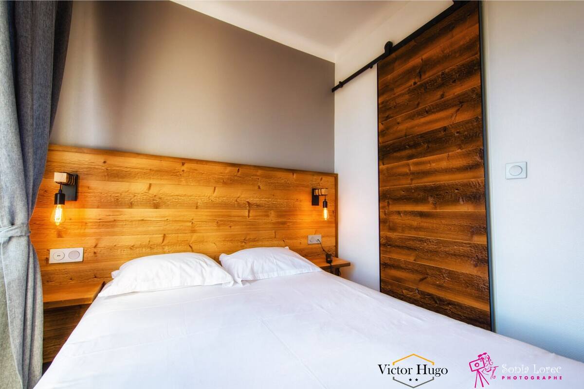 Habitación de hotel con cabecera de madera, dos lámparas de noche, cama con ropa blanca y puerta corredera de madera.