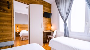 A room with two beds, a desk with a chair, a television on the wall, and a window with curtains. - Victor Hugo (Lorient)
