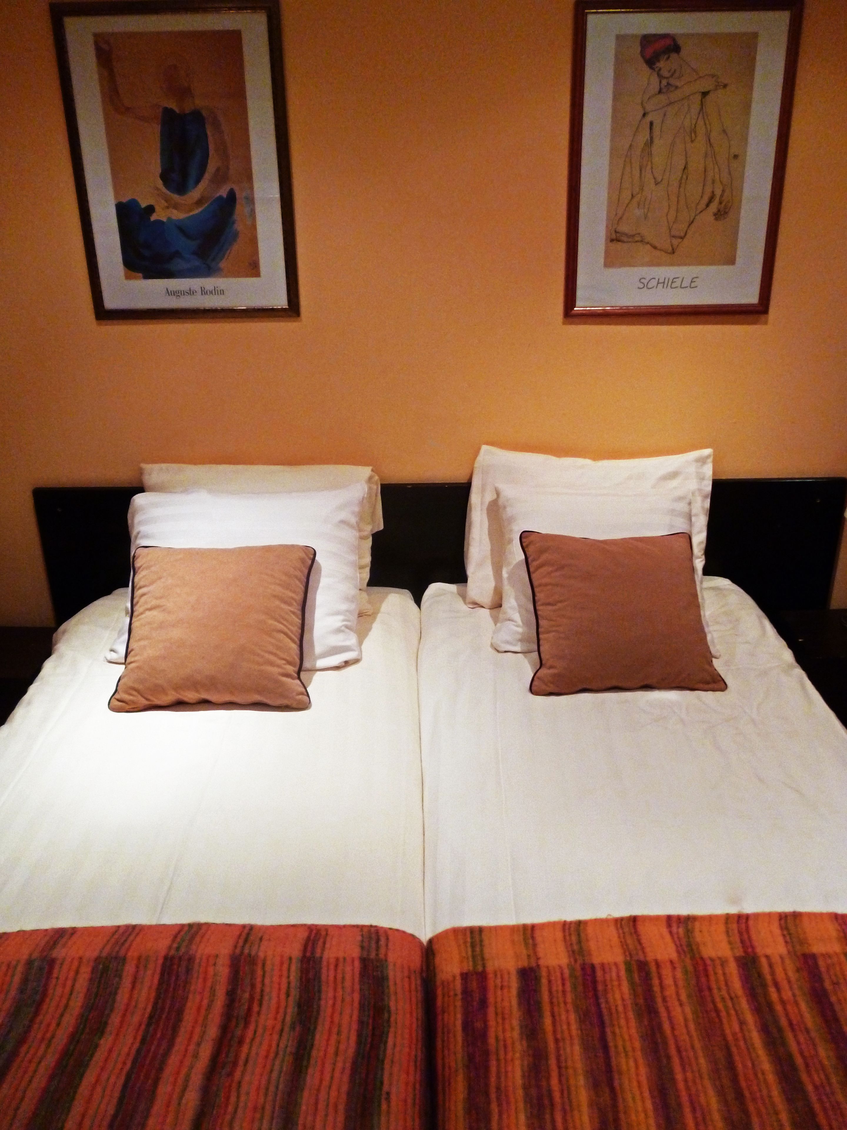 Uma cama de casal com lençóis brancos e duas almofadas marrons, uma obra de arte na parede acima e um cobertor listrado no pé da cama.